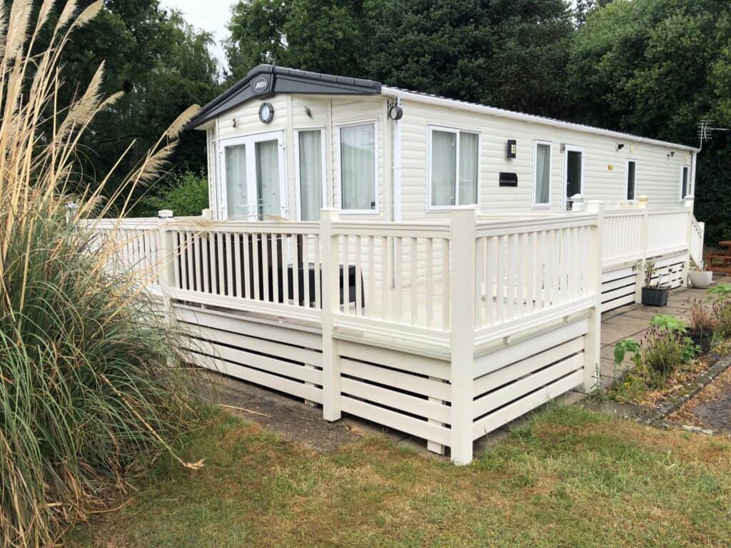 A white caravan with large windows and a wraparound veranda featuring white railings, surrounded by greenery and grass, with trees in the background. | Shorefield Caravan