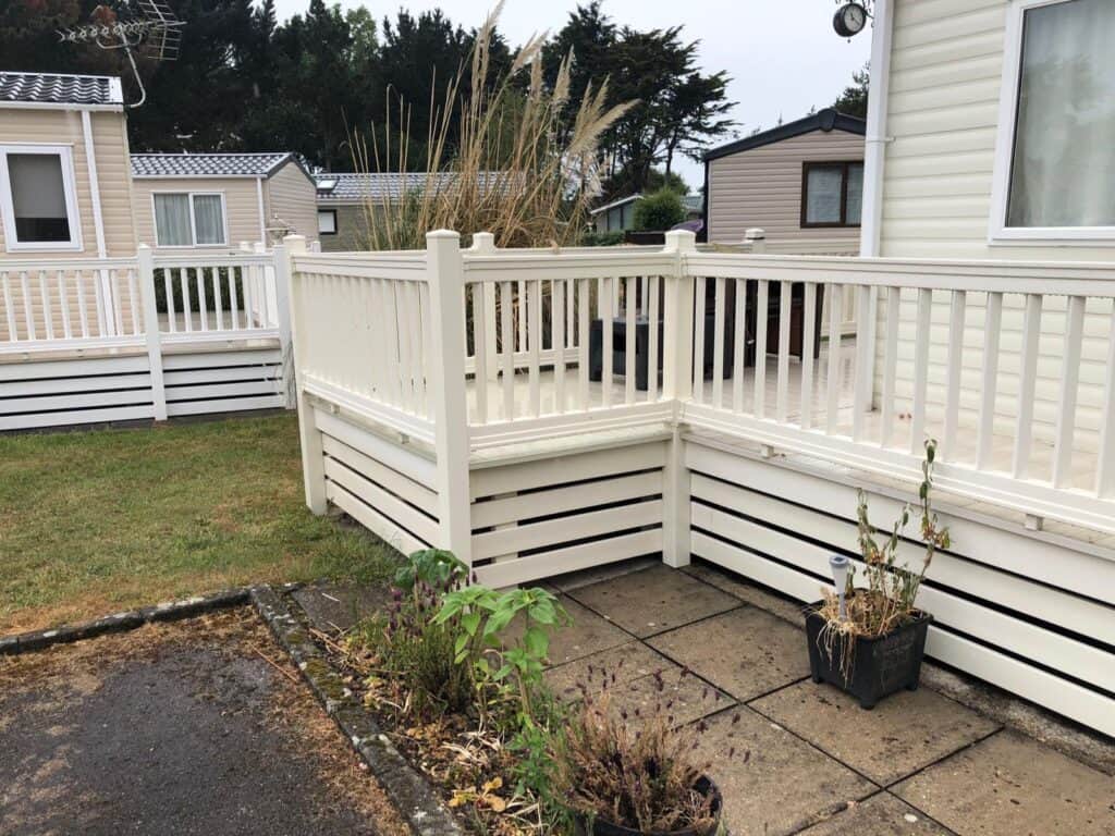 A wooden decking with white railings is attached to a mobile home. Two potted plants sit on a paved area, with grass and other mobile homes in the background under a cloudy sky. | Shorefield Caravan