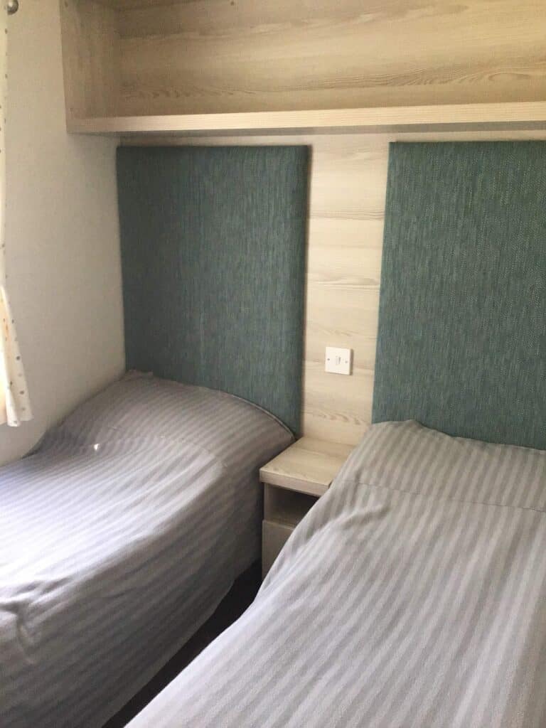 Two single beds with grey striped bedding are placed parallel with a small bedside table and power socket between them. Upholstered teal headboards and a wooden shelf are mounted on the wall above the beds. | Shorefield Caravan