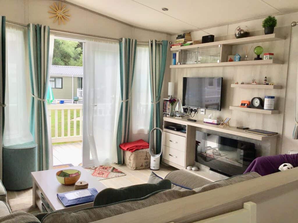 Bright living room with large windows, teal curtains, a TV on a light wood media unit, shelves with decor, a fireplace, a sofa, coffee table, and an outside view of a porch and green area. | Shorefield Caravan