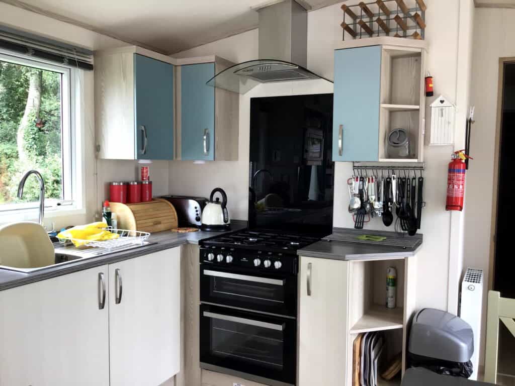 Modern kitchen with blue and white cupboards, a black oven and hob, utensils hanging on the wall, crockery drying by the sink under a window, a kettle, and a fire extinguisher on the side wall. | Shorefield Caravan