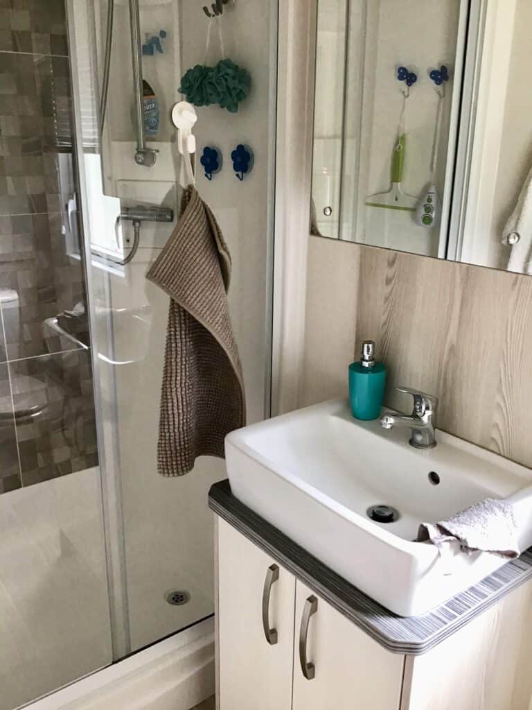 A modern bathroom with a glass shower, beige towel hanging on a hook, white basin with a teal soap dispenser, and a grey flannel on the counter. Blue shower puffs and toiletries are visible in the shower. | Shorefield Caravan