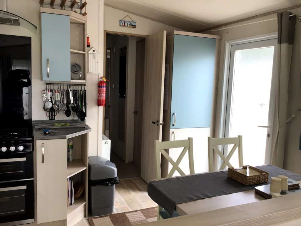 A modern kitchen and dining area with light wooden cupboards, a cooker, utensils hanging on a rack, a table with chairs, and an open door letting in natural light. A fire extinguisher is mounted on the wall. | Shorefield Caravan