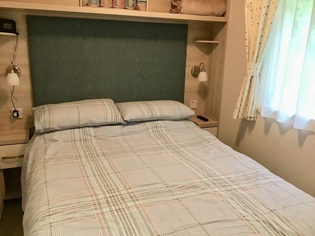 A neatly made bed with checked bedding, two pillows, a teal upholstered headboard, and built-in shelves on beige walls. Light from a window with white curtains brightens the cosy bedroom. | Shorefield Caravan
