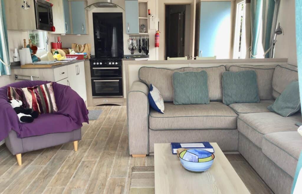 A cosy living space with a light grey sectional sofa, a purple armchair with soft toys and cushions, a modern kitchen area, wood-style flooring, and a coffee table with bowls and magazines. | Shorefield Caravan