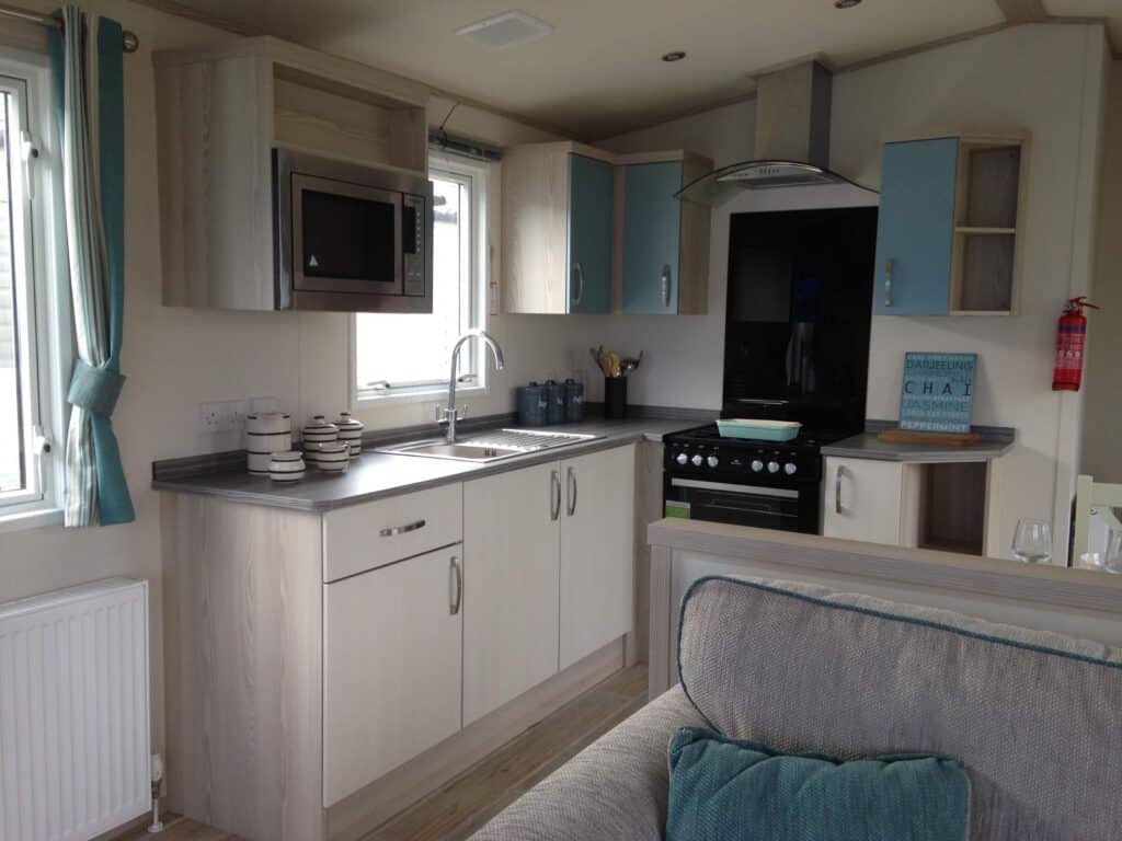 Modern kitchen with light wood cupboards, grey worktops, a built-in microwave, cooker with oven, sink under a window, blue accents, and a fire extinguisher on the wall. Sofa with blue cushions in the foreground. | Shorefield Caravan