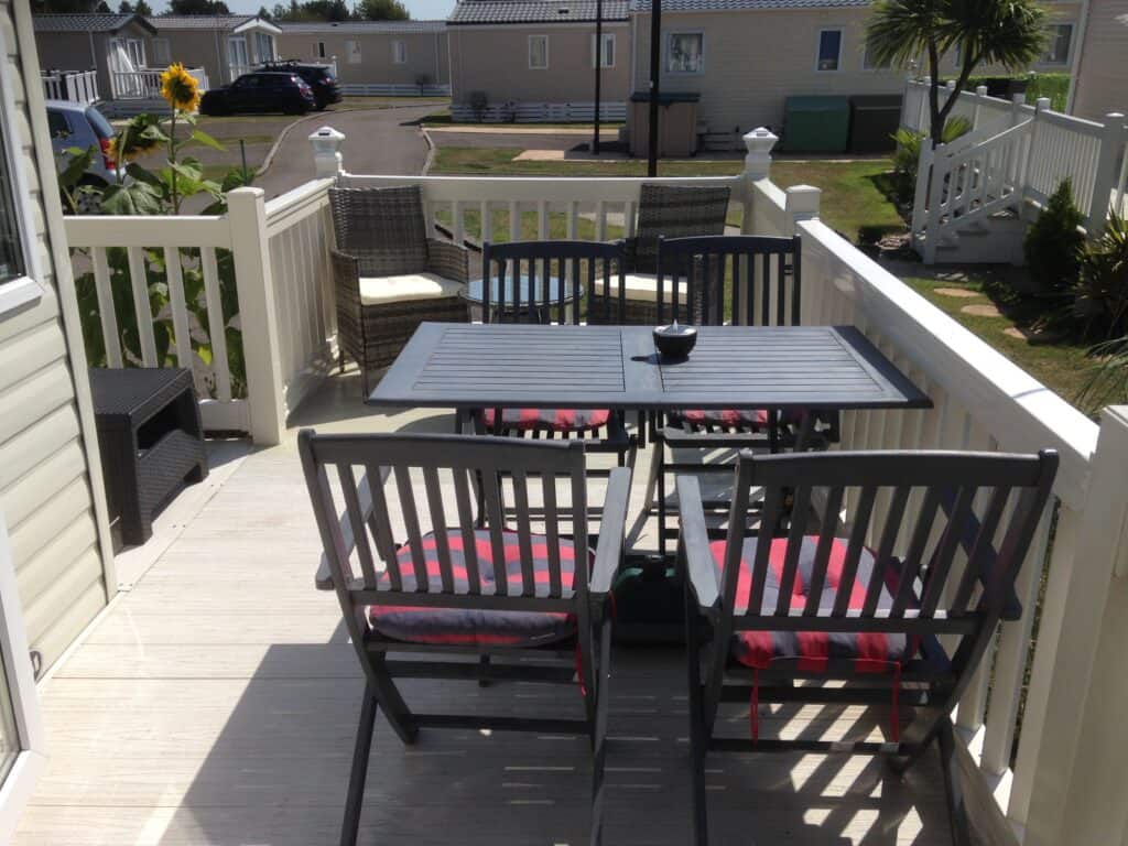 A patio with a table and four chairs with striped cushions, set on a wooden decking outside a caravan. Sunflowers and other plants are visible, with more caravans in the background. | Shorefield Caravan